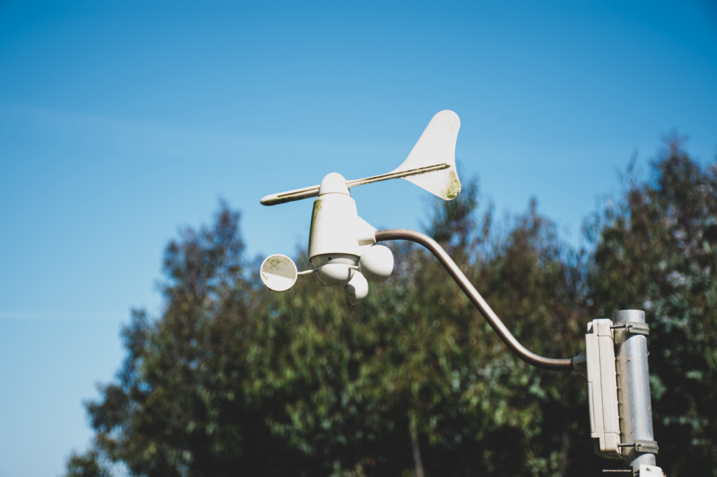 A white anemometer measuring wind speed against a clear blue sky, surrounded by greenery.