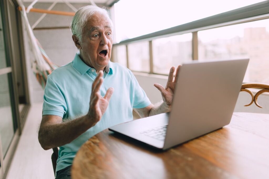 Elderly man looking shocked while using a laptop on a balcony, depicting excitement or surprise.
